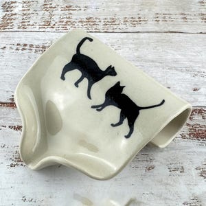 May include: A cream-coloured ceramic spoon rest with a curved, concave shape and a smooth, glossy finish. The surface features two black cat silhouettes, one standing and one in a playful pose, positioned centrally on the off-white background.