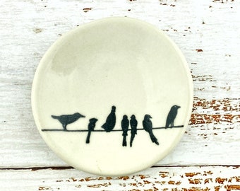 May include: A small, round, off-white ceramic dish featuring a black graphic illustration of seven birds perched on a single horizontal line. The surface of the dish is smooth and glossy, with a slightly irregular, hand-formed circular rim.