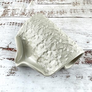 Peut inclure: Un porte-savon en céramique gris clair orné d'un motif en relief représentant de petites tortues de mer sur toute sa surface. L'objet présente une forme incurvée et inclinée avec un centre creux et une finition lisse et brillante.