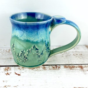 May include: A handcrafted ceramic mug with a forest design. The mug has a colour gradient, shifting from dark blue at the rim to turquoise and green at the base. The handle is green. The forest design is embossed on the mug.