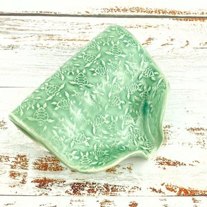 May include: A seafoam green ceramic dish with a raised turtle pattern. The dish has a wavy, irregular shape and is set against a white, distressed wood background. The turtles are evenly spaced across the surface.