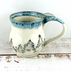 May include: A handmade ceramic mug with a cream-coloured body and a blue rim. The mug features a forest design in shades of grey and black. The handle has a unique design element. The mug is approximately 10 cm tall.