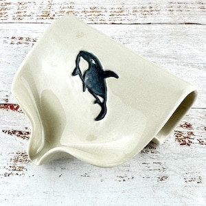 May include: A cream-coloured ceramic soap dish with a curved, ergonomic shape and a built-in drainage spout. The centre of the dish features a dark, hand-painted silhouette of an orca whale, providing a clear contrast against the smooth glaze.