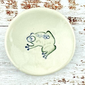Peut inclure: Petite assiette en céramique blanc cassé avec un motif de grenouille peint à la main. La grenouille est verte avec des détails bleus et a un aspect fantaisiste et caricatural. L'assiette est ronde, environ 10 cm de diamètre.