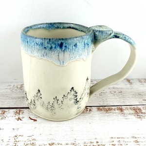 May include: A cream-coloured ceramic mug with a blue glazed rim. The mug features a forest scene with black tree silhouettes. The handle has a unique design with a small, decorative element. Ideal for tea or coffee, this mug is a handcrafted piece.