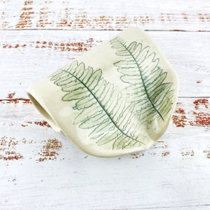 May include: A white ceramic dish with a fern leaf design. The dish is shaped like a half-circle and has a slightly raised edge.