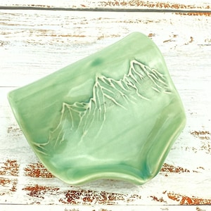 May include: A green ceramic dish with a raised design of mountains. The dish has a wavy edge and is ideal for holding jewellery or other small items.