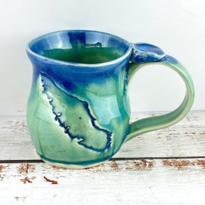 May include: A handcrafted ceramic mug with a distinctive design. The mug showcases a mix of blue and green glazes, producing a marbled appearance. The handle is curved, and the mug holds about 350 ml.