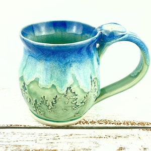 May include: A handcrafted ceramic mug with a distinctive design. The mug showcases a colour gradient, shifting from deep blue at the rim to turquoise and green at the base. The handle is a matching green. The mug has a textured, organic feel.
