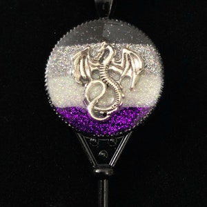 May include: A silver dragon charm pendant with a black and white glitter background and a purple glitter bottom. The pendant is attached to a black cord necklace.