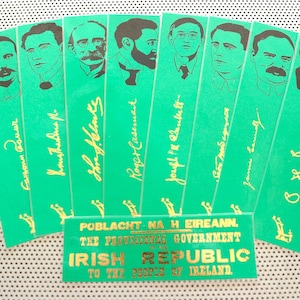 May include: A collection of green bookmarks featuring black portraits and gold signatures. The bookmarks are fanned out, with a small rectangular card at the base. The card displays gold text: "POBLACHT NA H EIREANN THE PROVISIONAL GOVERNMENT OF THE IRISH REPUBLIC TO THE PEOPLE OF IRELAND."