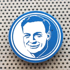 May include: A blue and white circular sticker with a white silhouette of a man's face. The man is smiling and has short, dark hair.