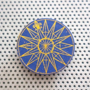 Peut inclure: Une boussole bleue et dorée avec un motif de fleur de lys. La boussole est sur un fond blanc avec un motif de petits trous.