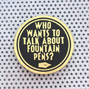 Peut inclure: Un bouton rond noir et or avec le texte "WHO WANTS TO TALK ABOUT FOUNTAIN PENS?" en lettres jaunes. Une flèche pointe vers la droite.