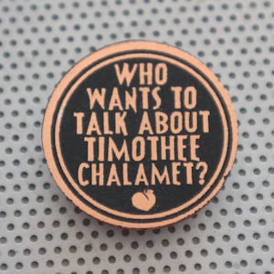 Puede incluir: Un botón redondo de color negro y melocotón con el texto "WHO WANTS TO TALK ABOUT TIMOTHEE CHALAMET?" en blanco.
