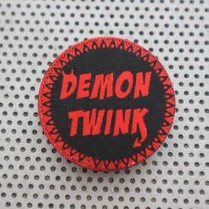 May include: A red and black round button with the text "DEMON TWINK" in red letters.