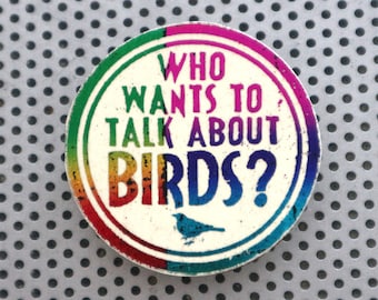 Pode incluir: Um botão redondo e colorido com o texto "WHO WANTS TO TALK ABOUT BIRDS?" em uma fonte extravagante. O botão tem uma borda de arco-íris e uma pequena ilustração de um pássaro azul. O fundo é uma superfície de metal perfurada.