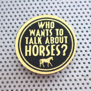 May include: A circular wooden badge featuring the phrase "WHO WANTS TO TALK ABOUT HORSES?" in yellow text on a black backdrop. A yellow horse silhouette is below the text. The badge is set against a grey, perforated metal surface.