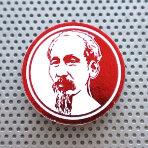 Puede incluir: Un botón circular rojo con una imagen blanca de un hombre con barba y bigote. El hombre mira hacia la derecha.