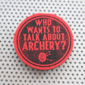 May include: Red and black round button with the text "WHO WANTS TO TALK ABOUT ARCHERY?" in white lettering. A black target with three arrows is in the center of the button.