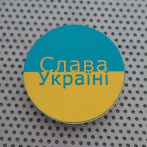 May include: A round sticker with the Ukrainian flag colors, blue and yellow. The sticker has the text "Слава Україні" in black lettering.