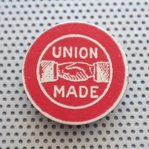 Op de afbeelding: Een ronde rode knop met witte tekst die "UNION MADE" luidt en een wit beeld van twee handen die elkaar schudden.