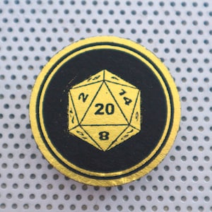May include: A gold-colored twenty-sided die with black numbers is displayed on a black and gold circular coaster. The coaster has a textured surface.