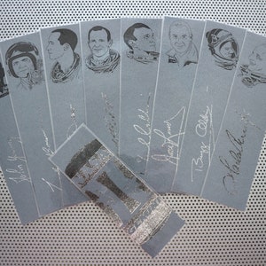 Puede incluir: Un conjunto de ocho marcapáginas plateados con ilustraciones de astronautas con sus firmas. Los marcapáginas están dispuestos en forma de abanico sobre un fondo gris.