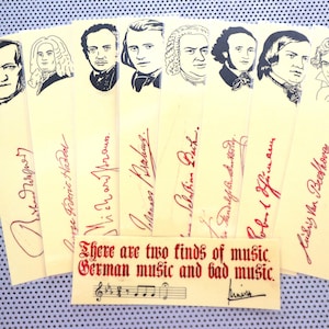 Puede incluir: Un conjunto de ocho marcapáginas amarillos con retratos de compositores famosos, incluyendo Johann Sebastian Bach, Ludwig van Beethoven y Wolfgang Amadeus Mozart. Cada marcapáginas incluye el nombre del compositor y su firma en tinta roja. Los marcapáginas están acompañados de una tarjeta amarilla más pequeña con el texto "Hay dos tipos de música. Música alemana y mala música." y un pentagrama musical con notas.