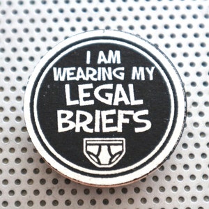 May include: A black and white round button with the text "I am wearing my legal briefs" and a white outline of underwear.