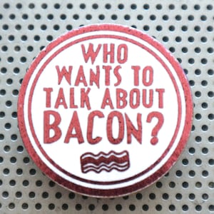 Puede incluir: Un botón redondo con fondo blanco y borde rojo. El texto "WHO WANTS TO TALK ABOUT BACON?" está en rojo, con un gráfico de tocino en la parte inferior. El botón está sobre una superficie plateada perforada.