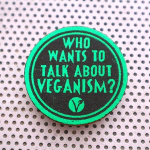 May include: A circular button with a black centre and a green border. The text "WHO WANTS TO TALK ABOUT VEGANISM?" is printed in green. A green "V" symbol is at the bottom. The button is on a silver, perforated surface.