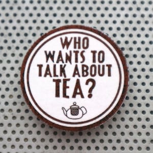 Puede incluir: Un botón redondo blanco con un borde marrón. El botón tiene el texto "WHO WANTS TO TALK ABOUT TEA?" en letras marrones. Una pequeña ilustración en blanco y negro de una tetera está debajo del texto.