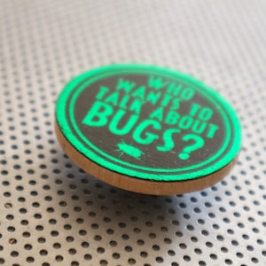 May include: A round, wooden button with a black background and green text that reads "Who wants to talk about bugs?"