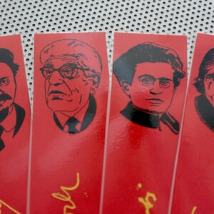 May include: Four red bookmarks with black and white illustrations of men's faces. Each bookmark has a gold signature at the bottom.