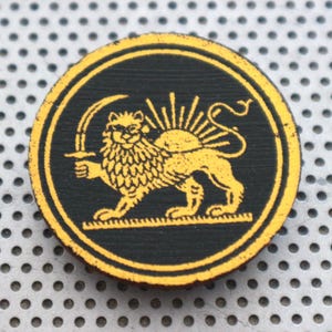 Puede incluir: Emblema redondo de madera con fondo negro y borde amarillo. Presenta un león amarillo sosteniendo una espada, con un diseño de rayos de sol detrás. El león está sobre una plataforma amarilla.