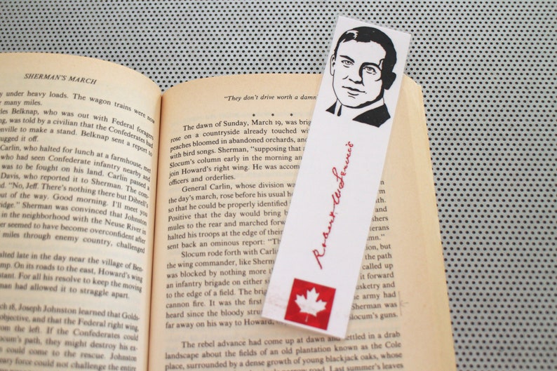 May include: A bookmark featuring a black and white illustration of a man with a mustache and a signature that reads "Robert E. Lee." The bookmark also features a red maple leaf on a white background.