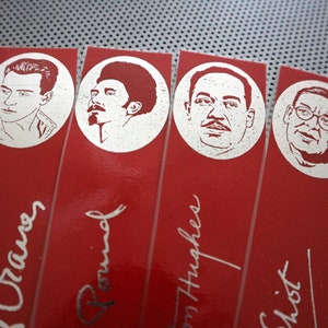 May include: A close-up of several red bookmarks with silver illustrations of faces in circles. The names of the people are written in cursive below the illustrations. The bookmarks are arranged diagonally.