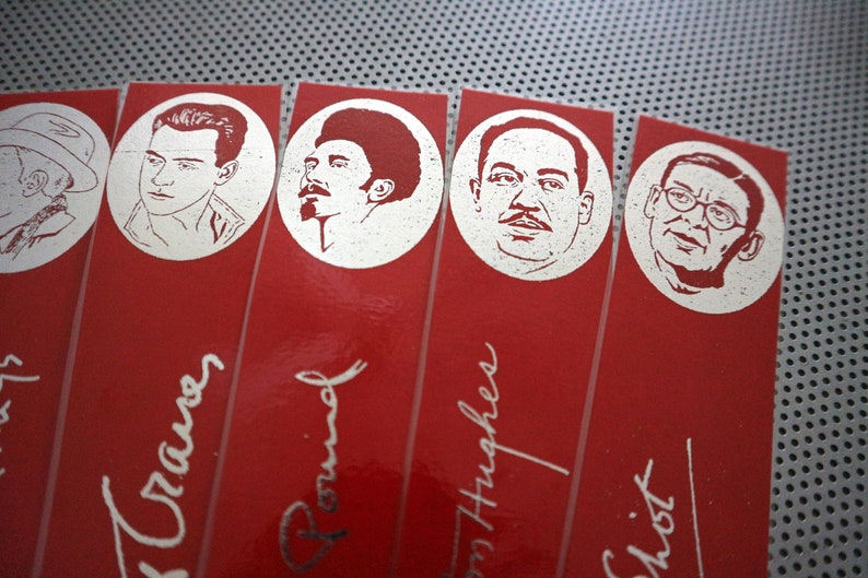 May include: A close-up of several red bookmarks with silver illustrations of faces in circles. The names of the people are written in cursive below the illustrations. The bookmarks are arranged diagonally.