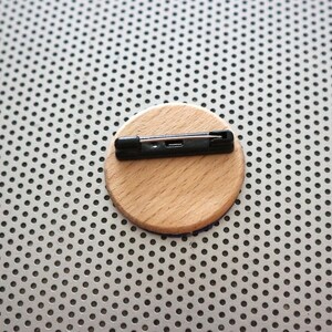May include: A round wooden pin back with a black metal pin attached. The pin back is on a silver background.