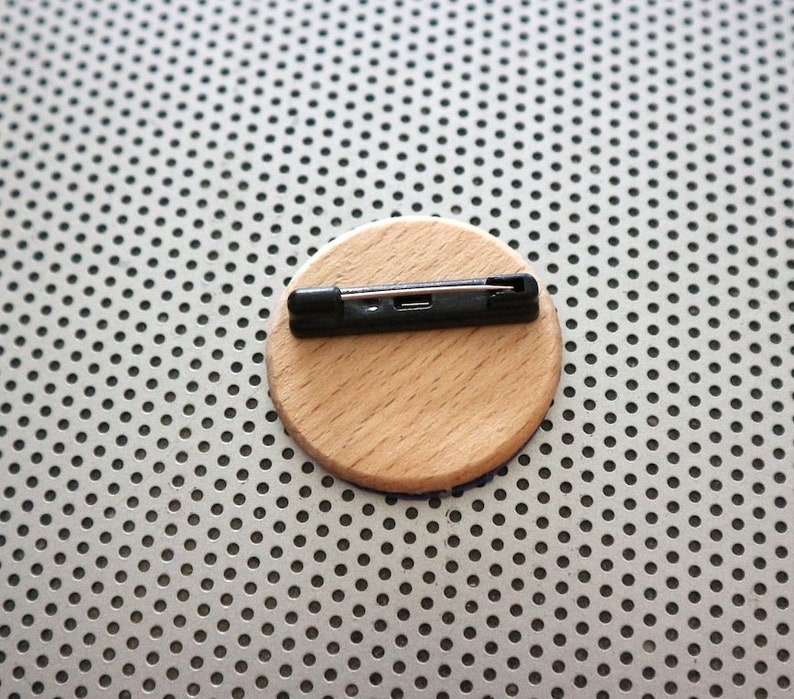 May include: A round wooden pin back with a black metal pin attached. The pin back is on a silver background.