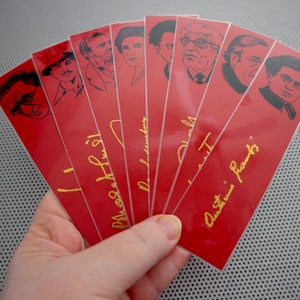 May include: Set of eight red bookmarks with black and white illustrations of famous authors. Each bookmark features a gold signature of the author, including "J.K. Rowling", "Maya Angelou", "Neil Gaiman", "Stephen King", "Jane Austen", "Charles Dickens", "Virginia Woolf", and "Ernest Hemingway".