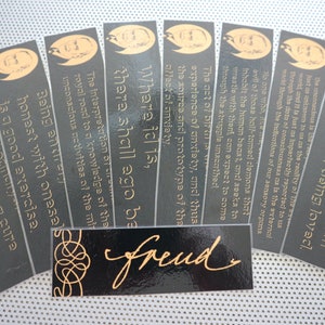 Puede incluir: Un conjunto de marcapáginas negros y dorados con citas de Sigmund Freud. Los marcapáginas presentan un retrato de Freud y su firma.