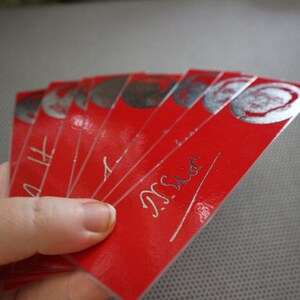 May include: A hand holds a fanned stack of red rectangular cards. Each card has a silver circular design at the top and silver text. The cards have a glossy finish. The text on the cards appears to read "N. Sist".