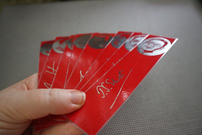 May include: A hand holds a fanned stack of red rectangular cards. Each card has a silver circular design at the top and silver text. The cards have a glossy finish. The text on the cards appears to read "N. Sist".