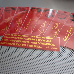 May include: A set of red bookmarks with gold lettering and images of people. The bookmarks feature quotes from famous people, including "Life is beautiful. Let the future generations cleanse it of all evil, oppression, and violence, and enjoy it to the full." - Leon Trotsky.