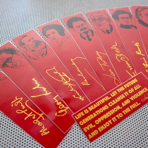May include: A set of red bookmarks with black and white illustrations of famous people. Each bookmark features a gold signature and a quote in gold lettering: "LIFE IS BEAUTIFUL LET THE FUTURE GENERATIONS CLEANSE IT OF ALL EVIL, OPPRESSION, AND VIOLENCE, AND ENJOY IT TO THE FULL."