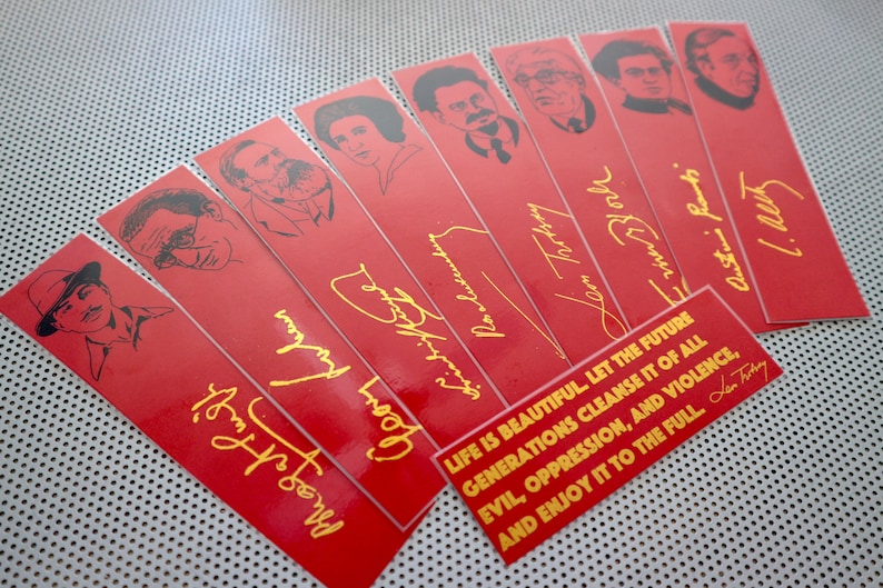 May include: A set of red bookmarks with black and white illustrations of famous people. Each bookmark features a gold signature and a quote in gold lettering: "LIFE IS BEAUTIFUL LET THE FUTURE GENERATIONS CLEANSE IT OF ALL EVIL, OPPRESSION, AND VIOLENCE, AND ENJOY IT TO THE FULL."