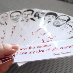 May include: A set of white bookmarks with black and white illustrations of people's faces. The bookmarks have red text that reads "I love this country. I love my idea of this country. - Gord Downie."