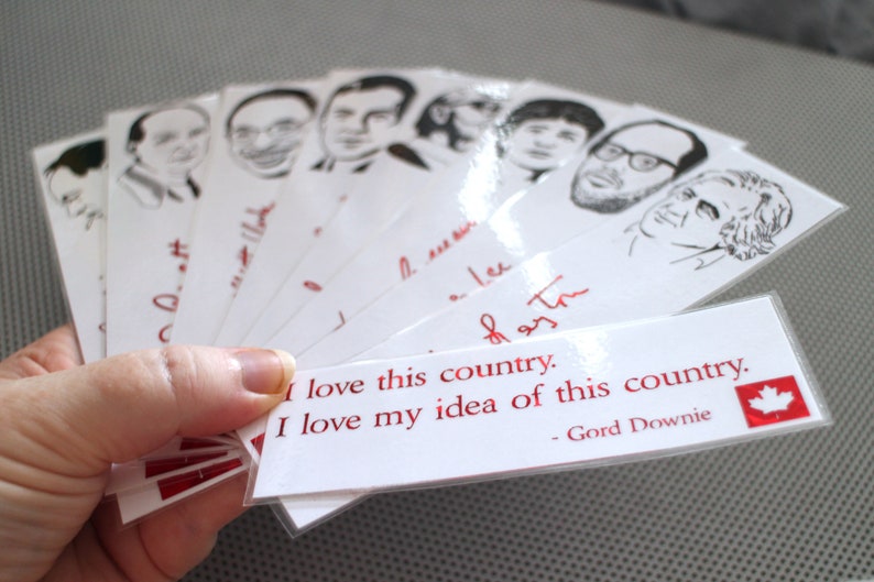 May include: A set of white bookmarks with black and white illustrations of people's faces. The bookmarks have red text that reads "I love this country. I love my idea of this country. - Gord Downie."